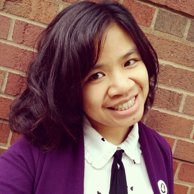 Selfie of Caitlan Truelove against a brick wall. She is smiling and wearing a white blouse with a knotted tie and a purple blazer.